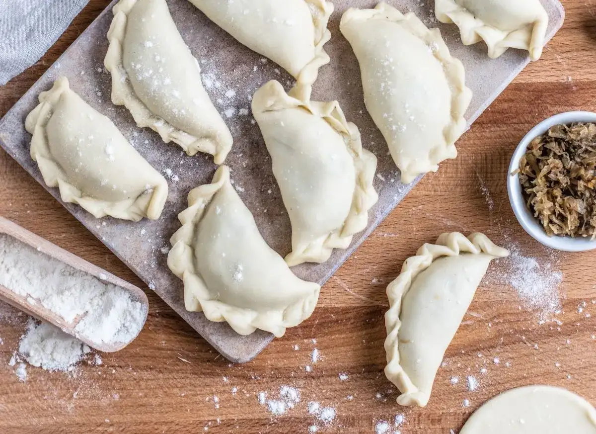 Surowe pierogi ruskie ułożone na desce, gotowe do zamrożenia. Obok miseczka z farszem i drewniana łopatka z mąką.