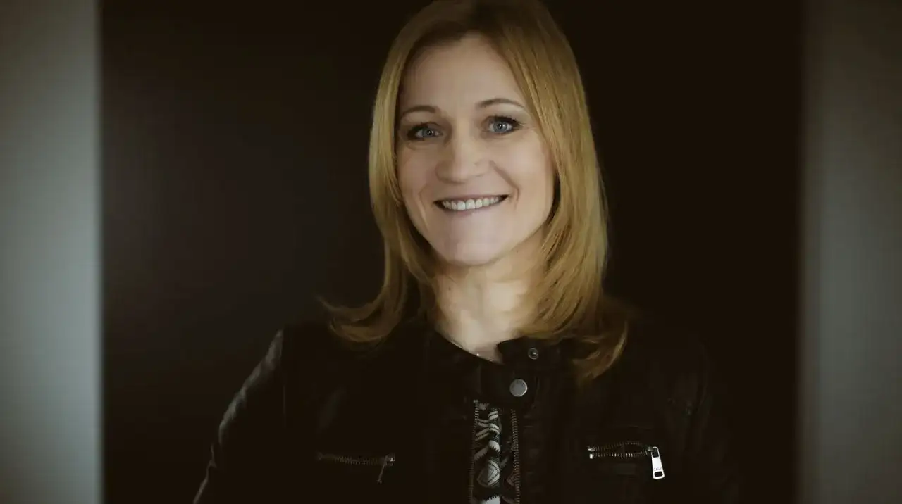 Uśmiechnięta blondynka w czarnej ramonesce na ciemnym tle.