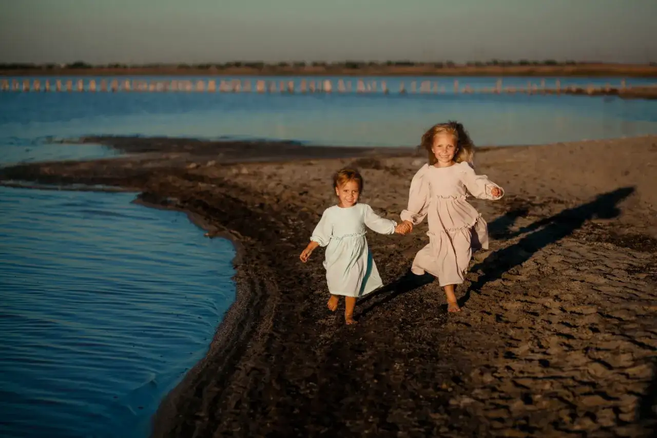 Dwie dziewczynki w sukienkach biegną trzymając się za ręce po piaszczystym brzegu nad wodą.