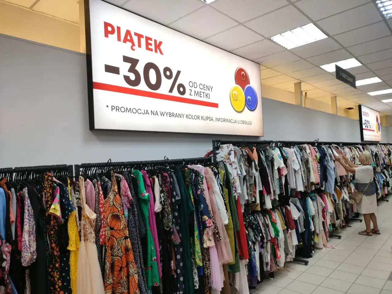 Wieszak z kolorowymi sukienkami i bluzkami w sklepie, baner z promocją -30%.