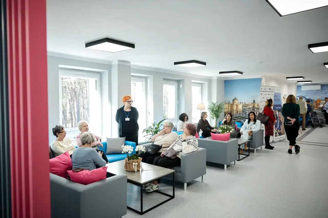 Grupa seniorów siedzi na kanapach w jasnym, nowoczesnym wnętrzu domu opieki.