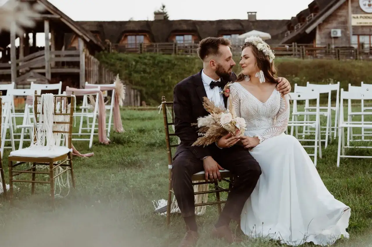 Para młoda siedzi na krzesłach na tle drewnianych budynków i białych krzeseł.