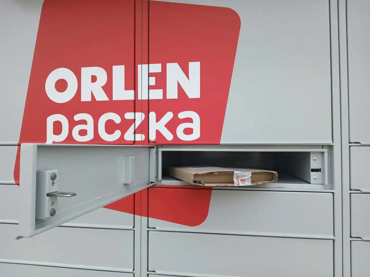 Szafka paczkomatu z logo Orlen Paczka i paczką w środku.