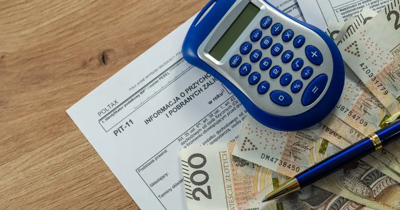Formularz PIT-11, kalkulator, długopis i polskie banknoty na drewnianym tle.