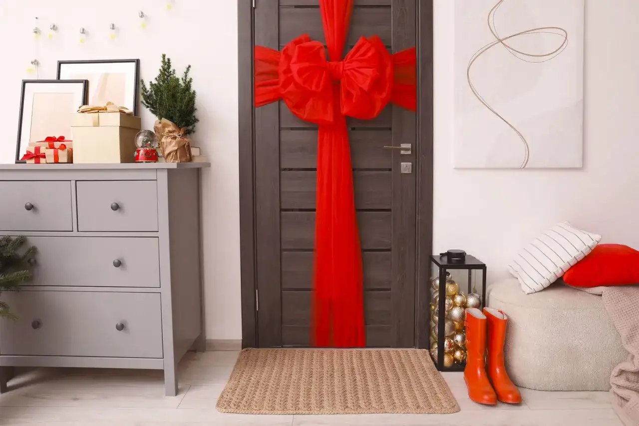 Jak zrobić kokardę na drzwi? Duża, czerwona kokarda z tiulu ozdabia ciemne drzwi, tworząc świąteczny nastrój. Obok stoją pomarańczowe kalosze.