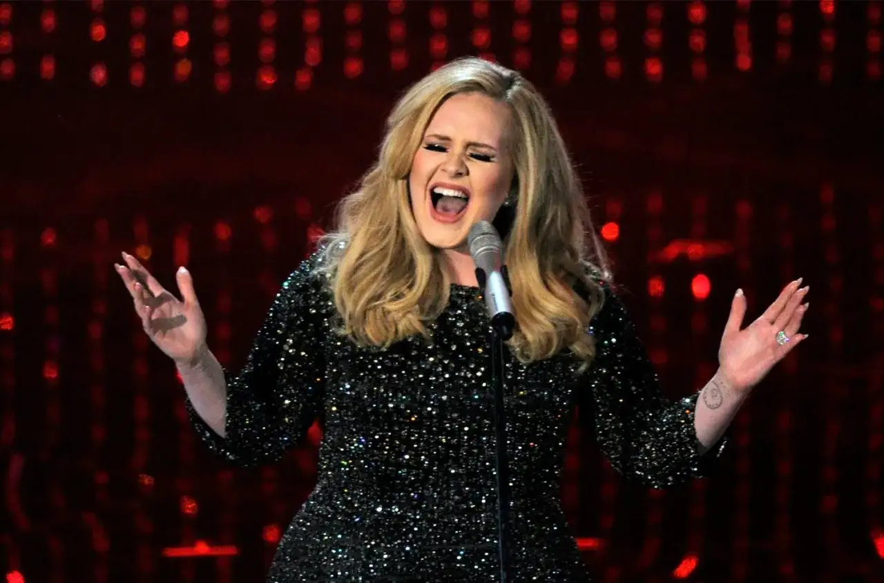 Brytyjska piosenkarka Adele śpiewa "Hello" w błyszczącej sukience, z emocjami na twarzy i uniesionymi rękami.