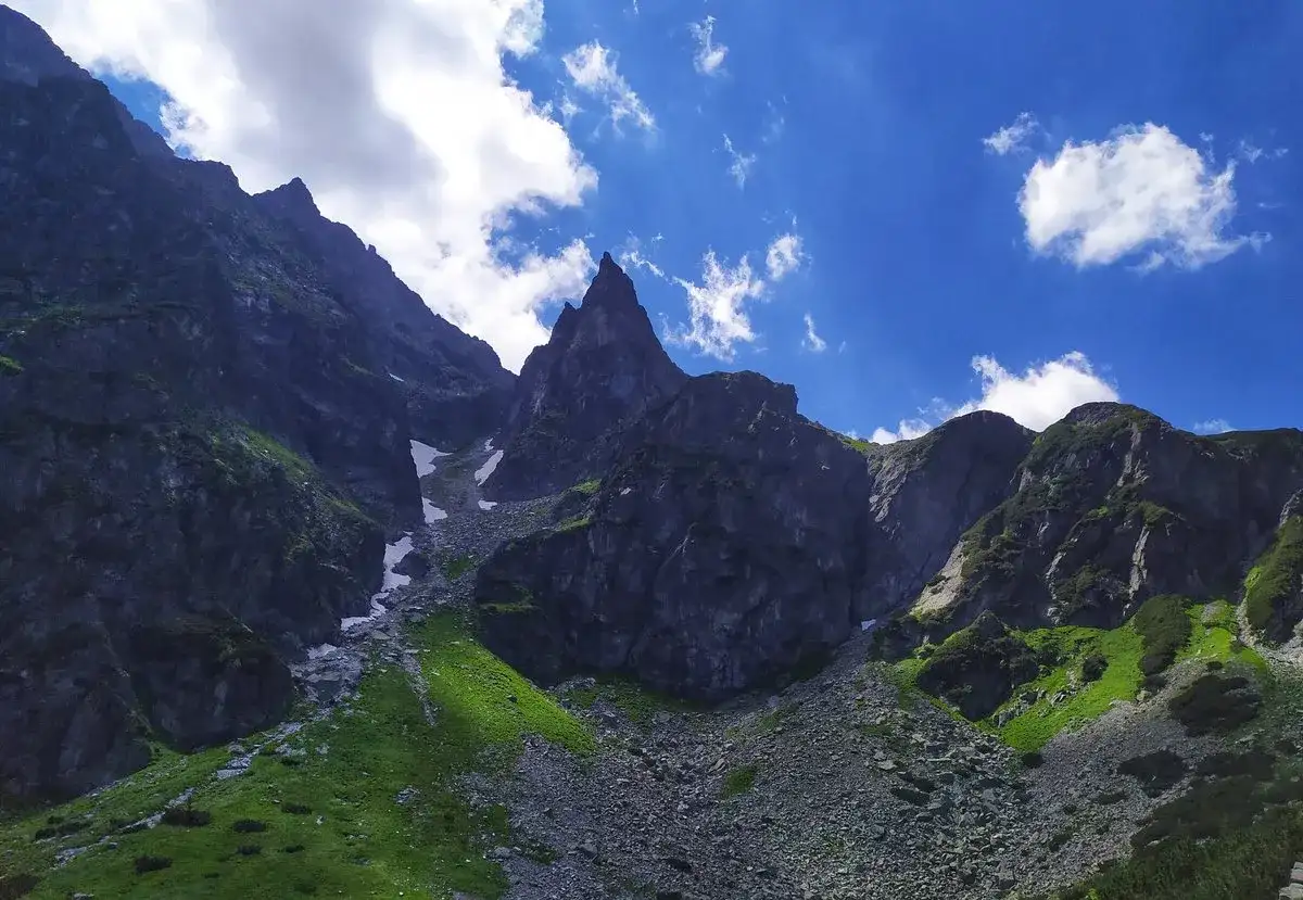 Kamieniste zbocza górskie z zieloną trawą i śniegiem pod błękitnym niebem z chmurami.