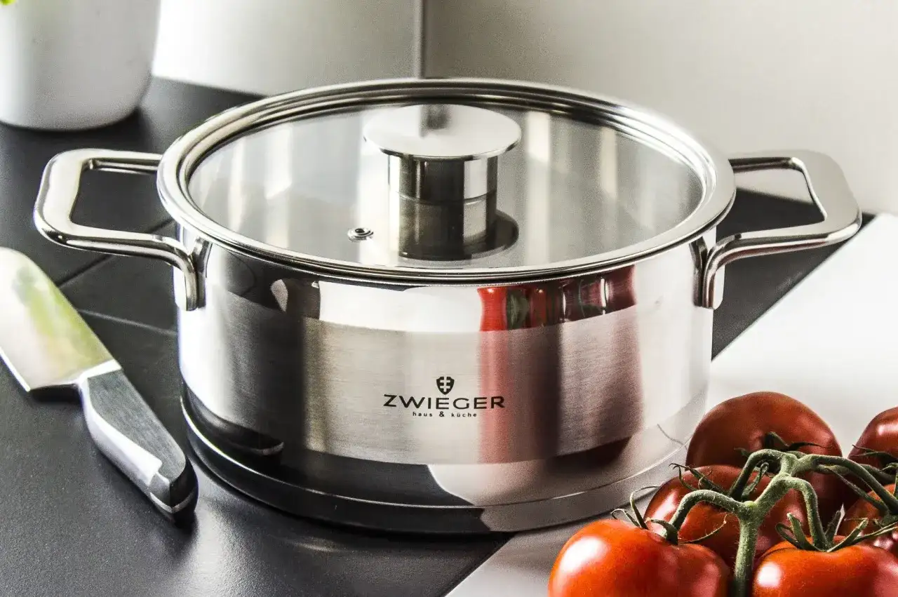 Stalowy garnek z logo Zwinger, idealny do przygotowania sosu pomidorowego. Obok nóż i świeże pomidory.