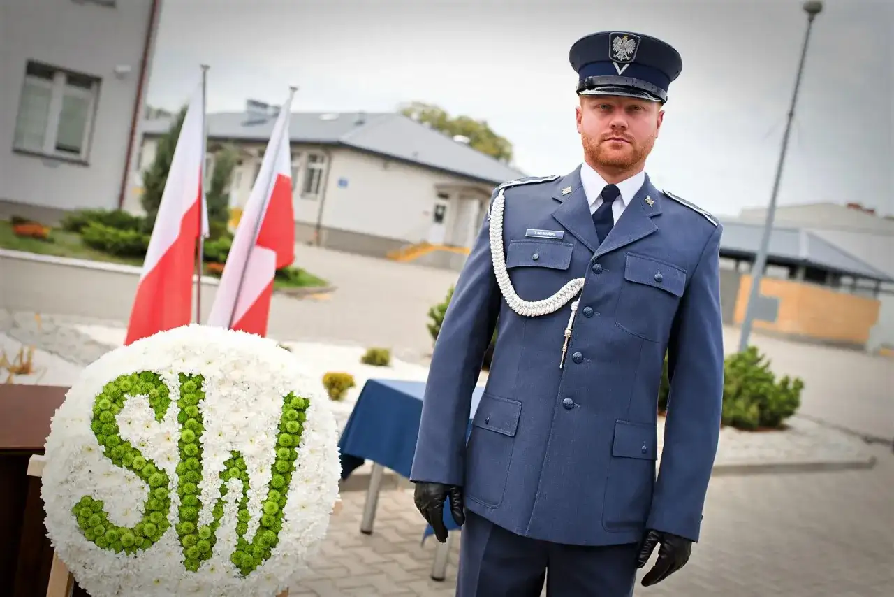 Funkcjonariusz Służby Więziennej w mundurze, obok wieńca z napisem SW i flag Polski.