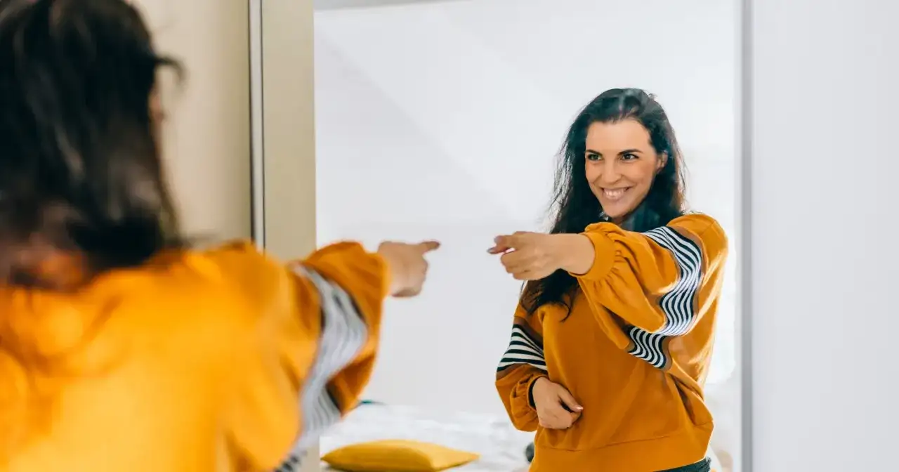 Uśmiechnięta kobieta w musztardowej bluzie wskazuje palcem na swoje odbicie w lustrze, emanując charyzmą.