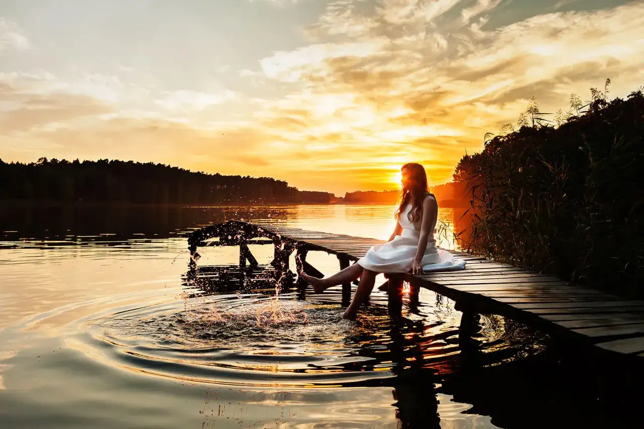 Dziewczyna w białej sukience na pomoście, pluska nogami w jeziorze podczas sesji urodzinowej w plenerze. Zachód słońca.