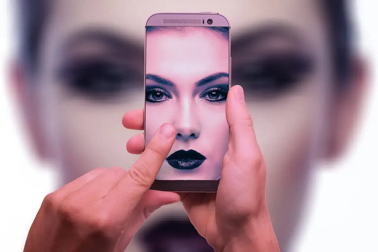 Dłonie trzymają smartfon, na ekranie którego widać twarz kobiety z mocnym makijażem. To przykład zaawansowanej fotografia obliczeniowa.