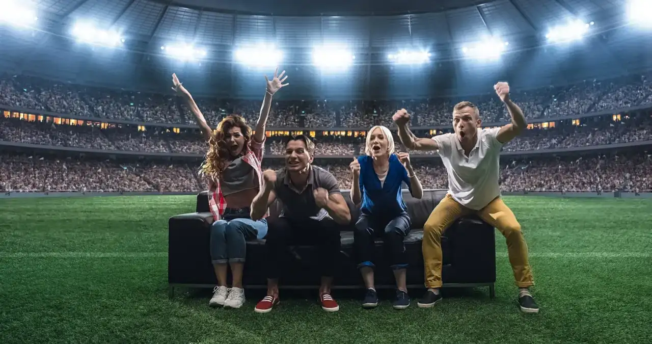 Cztery osoby siedzące na kanapie na boisku piłkarskim, kibicujące na tle stadionu.