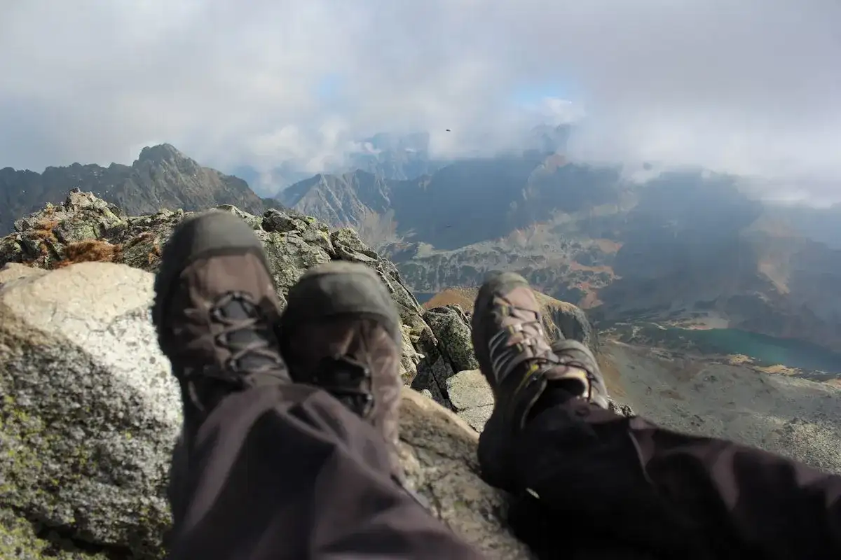 Stopy w butach trekkingowych na tle jesiennych Tatr. Październikowe słońce przebija się przez chmury, oświetlając skaliste szczyty i turkusowe jezioro.