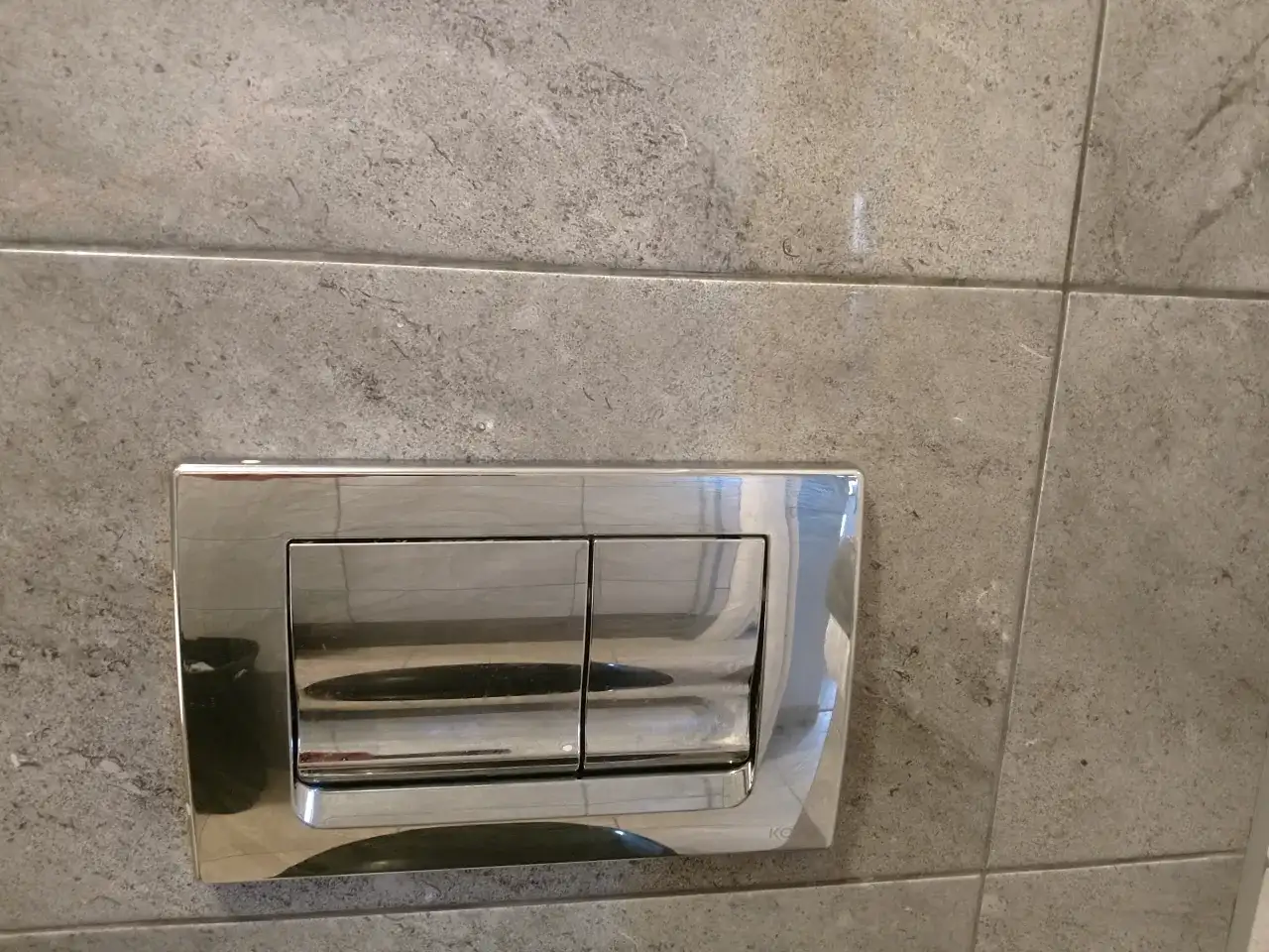 Chromowany przycisk spłuczki podtynkowej na szarej płytce. Dowiedz się, jak zdemontować przycisk spłuczki podtynkowej.