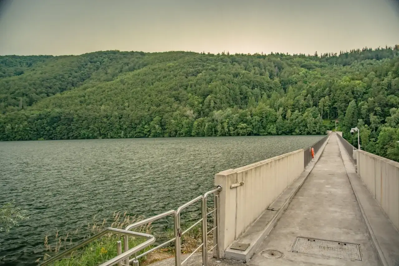 Zapora wodna z betonowym chodnikiem i balustradą, otoczona zielonymi wzgórzami i jeziorem.