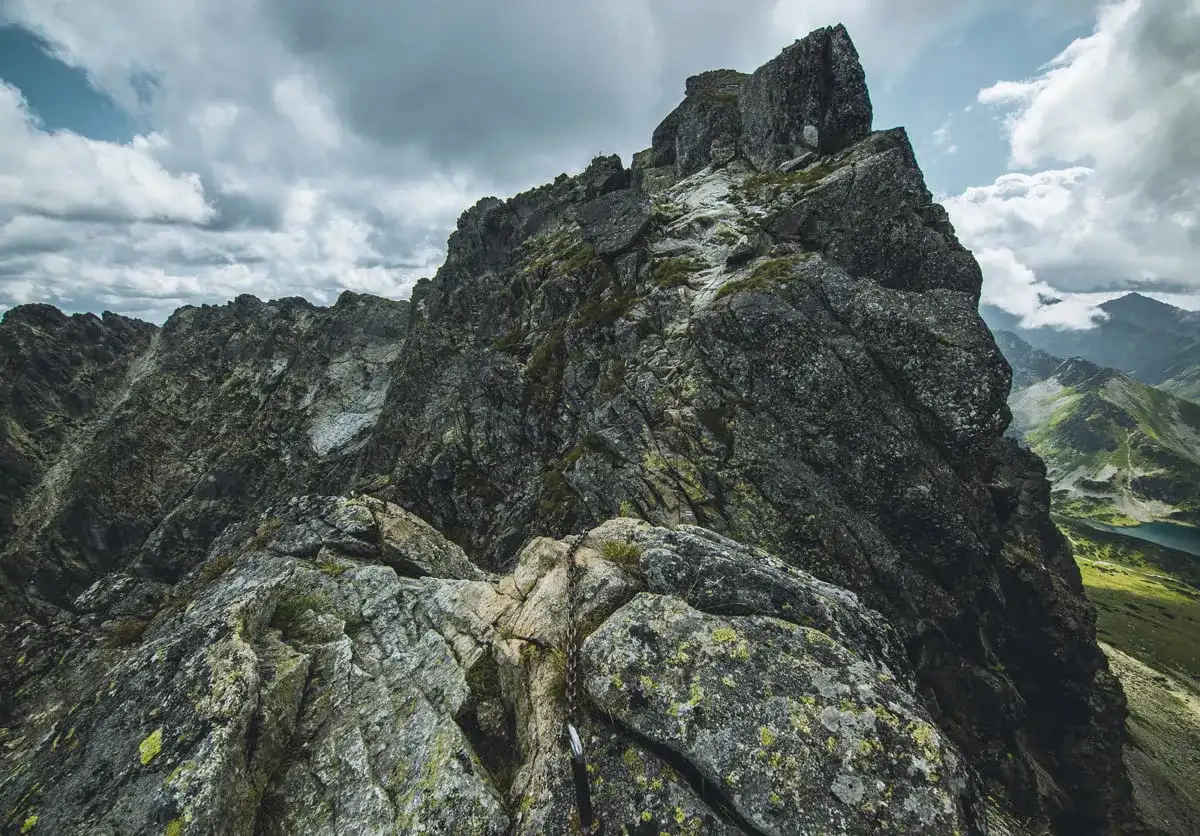 Kamienista trasa na Kozi Wierch, z łańcuchami ułatwiającymi wspinaczkę. Dramatyczne chmury nad skalistymi szczytami Tatr.