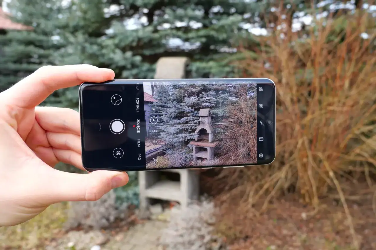 Dłoń trzyma smartfon z włączonym aparatem, kadrujący grill ogrodowy.