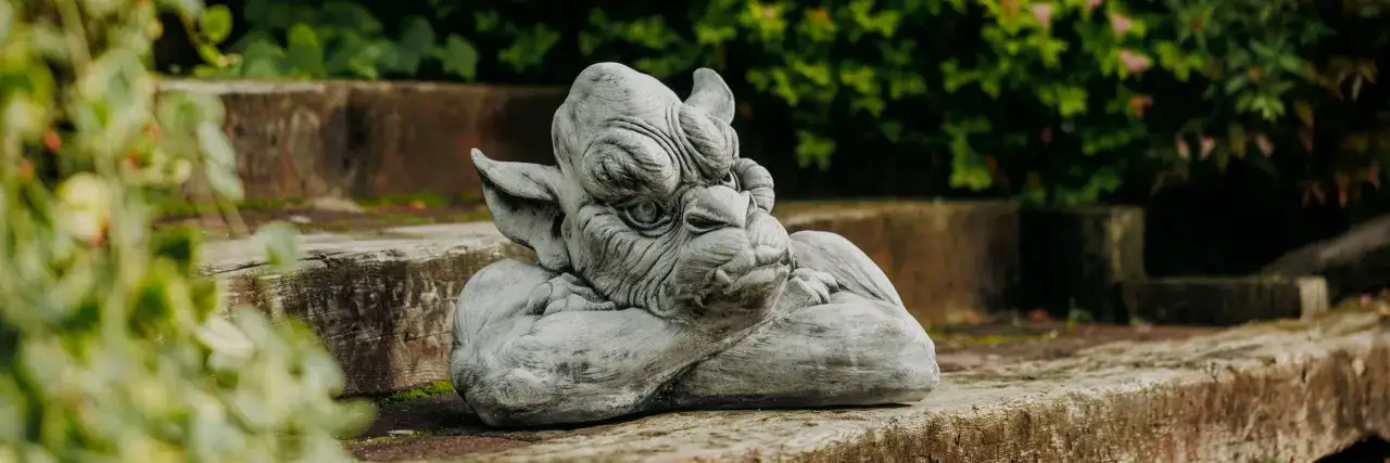 Graue Gartenskulptur eines Fabelwesens, das auf einem Holzstamm ruht, mit grünem Laub im Hintergrund.
