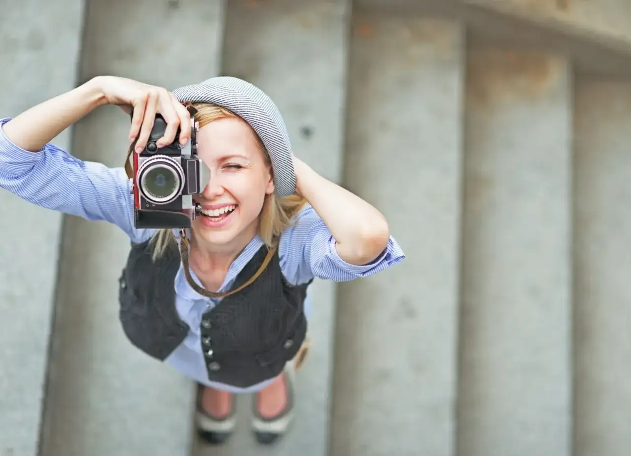 Uśmiechnięta dziewczyna w kapeluszu robi zdjęcie starym aparatem. To świetny pomysł na szkolenie z fotografii!