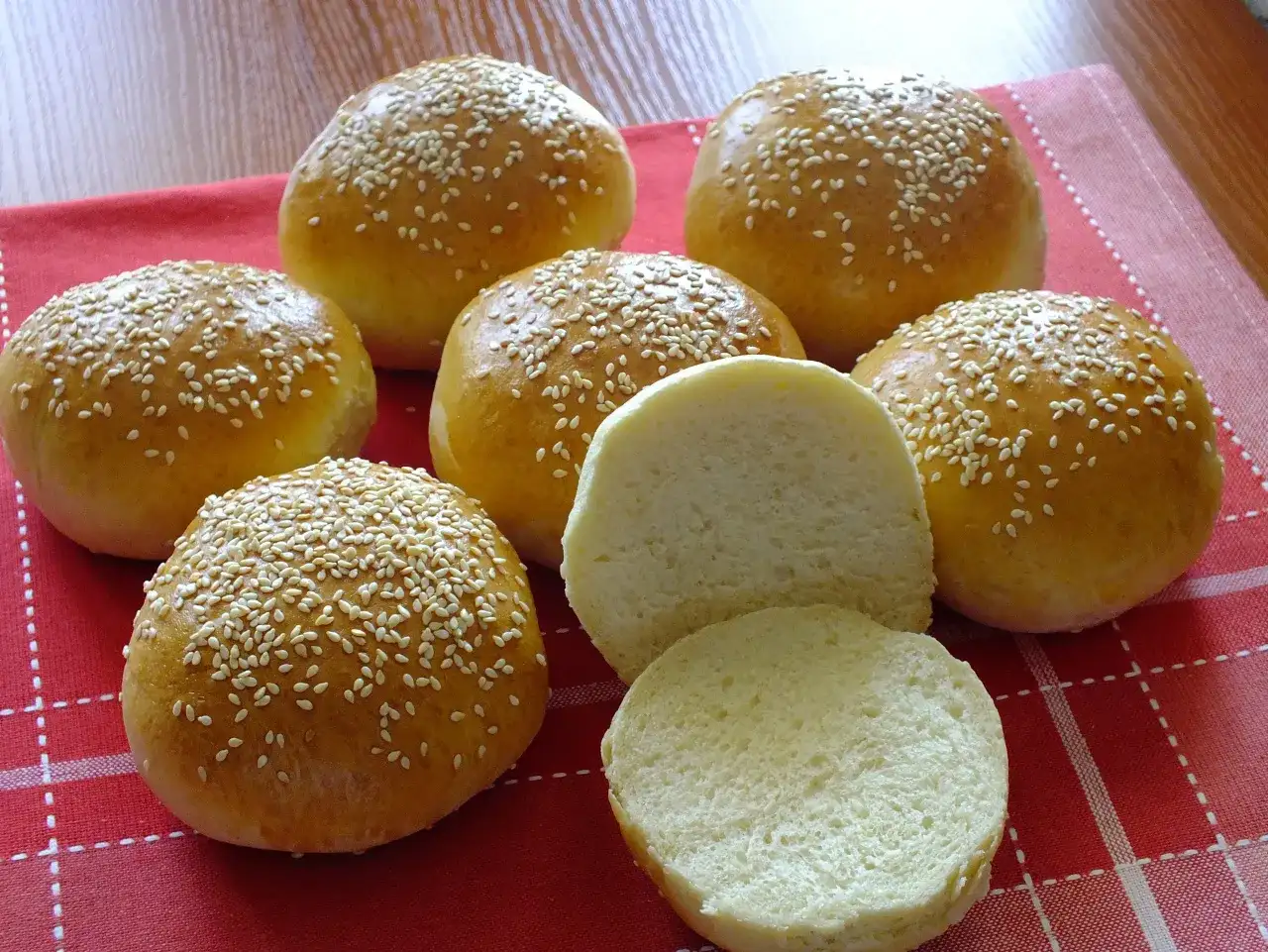 Kilka złotych bułek do burgerów posypanych sezamem, jedna przekrojona na pół.