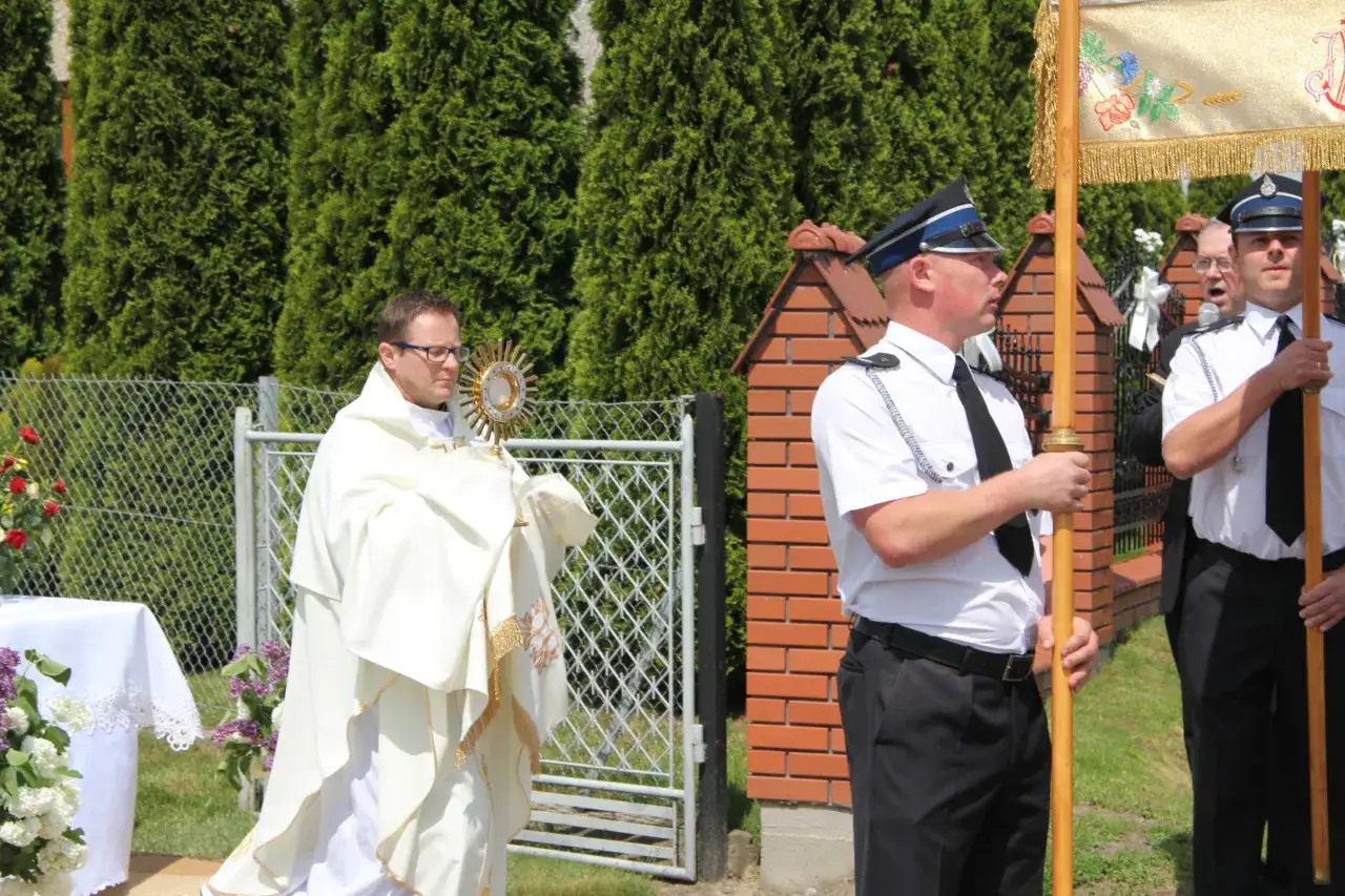 Ksiądz w białej szacie niesie monstrancję, obok stoją mężczyźni w mundurach.