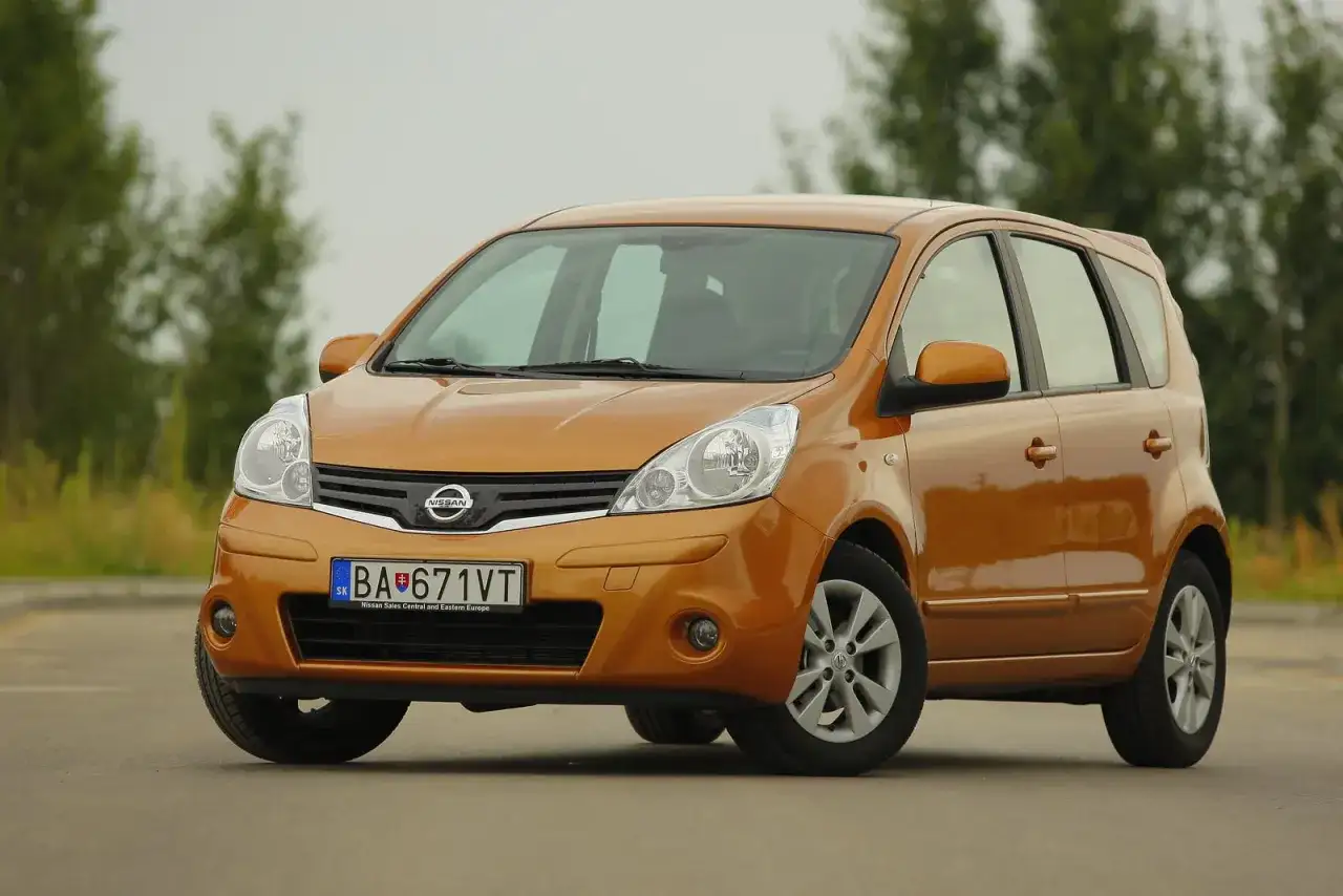 Pomarańczowy Nissan Note z przodu, na tle drzew.