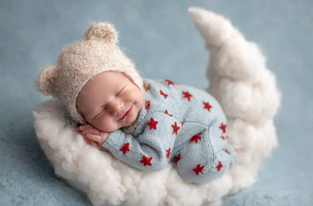 Uśmiechnięte niemowlę w czapce z uszkami śpi na chmurce w kształcie księżyca.