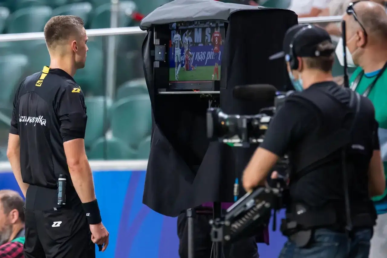 Sędzia obserwuje powtórkę na monitorze podczas meczu piłki nożnej. W tle operator kamery.
