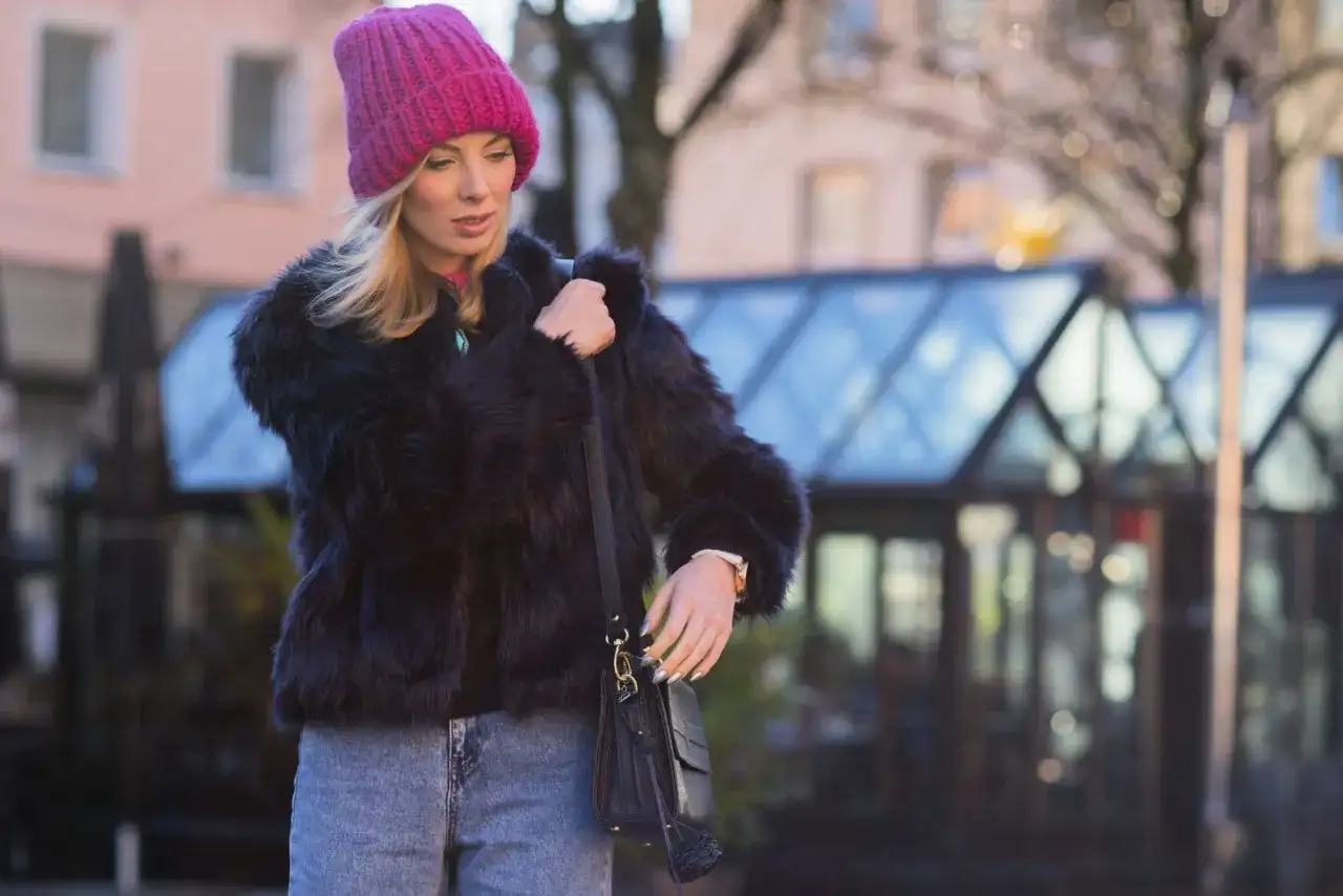Krótkie futerko stylizacje: blondynka w różowej czapce i czarnym futerku, z jeansami i torebką, na tle miasta.