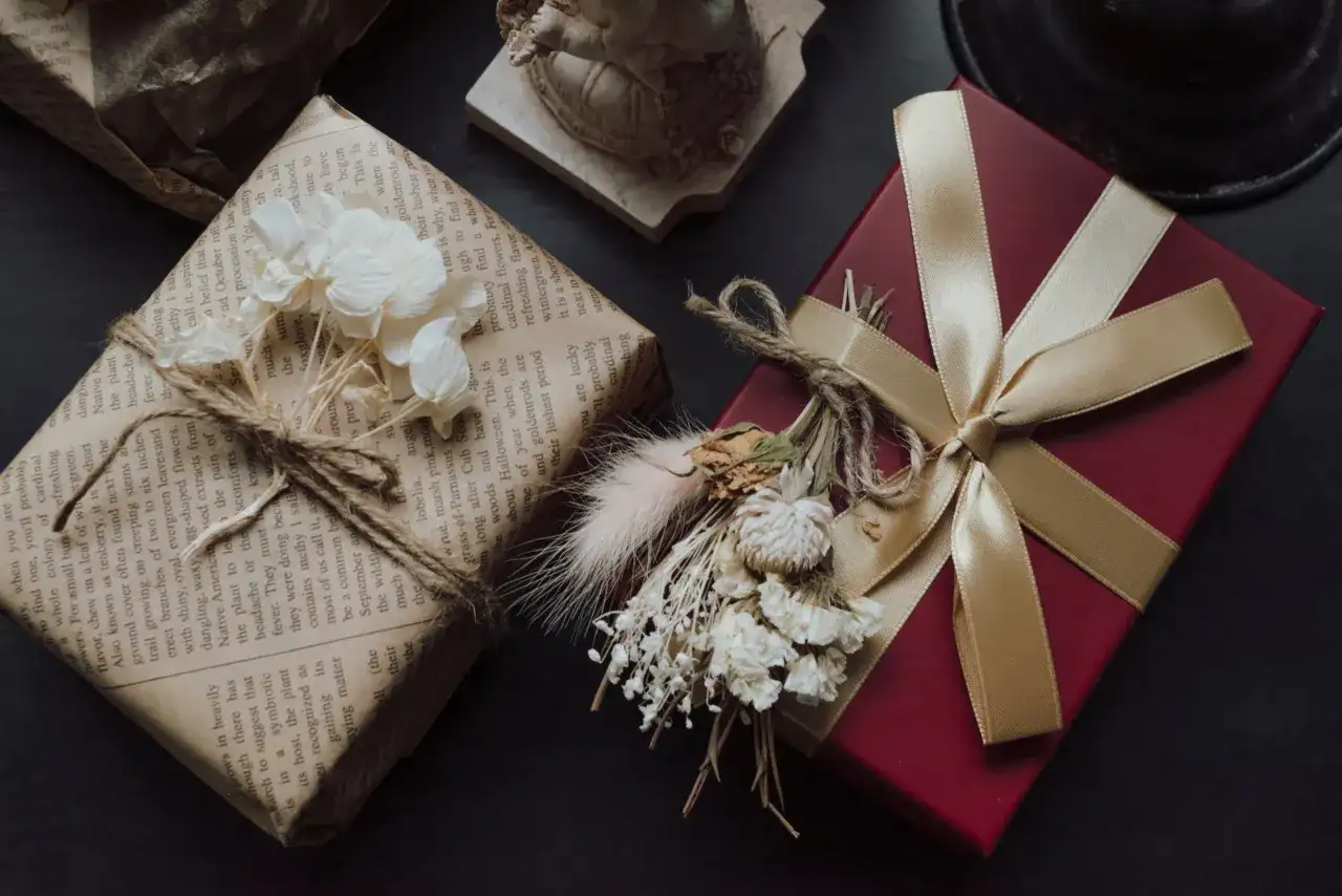 Elegancko zapakowane prezenty na urodziny: czerwone pudełko ze złotą wstążką i bukiecikiem suszonych kwiatów oraz prezent owinięty w papier z tekstem, ozdobiony sznurkiem i białymi kwiatami.