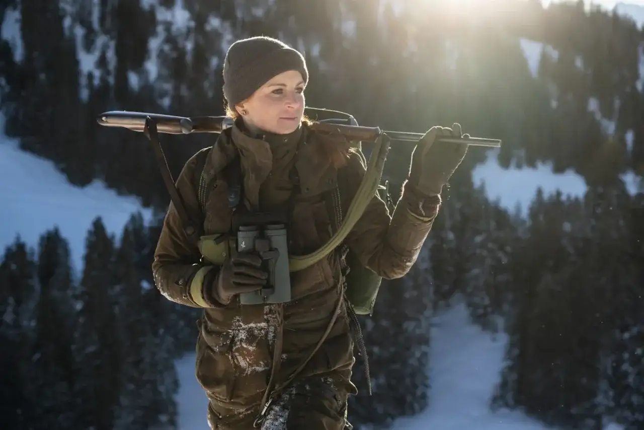 Kobieta w zimowym krajobrazie z karabinem na ramieniu i lornetką w dłoni. Czeka na to, co ustrzelił myśliwy.