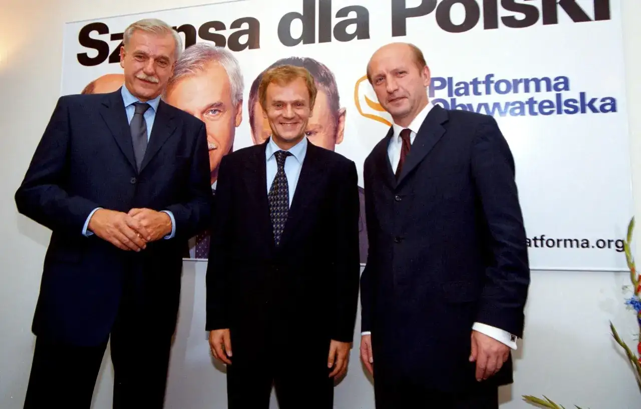Trzech mężczyzn w garniturach, założyciele Platformy Obywatelskiej, pozuje na tle banera z napisem "Szansa dla Polski".