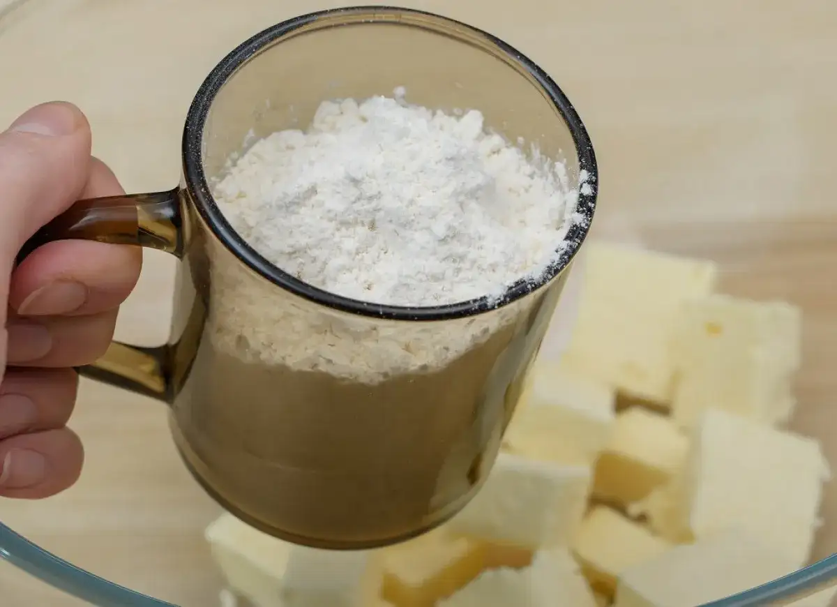 Dłoń trzyma kubek z mąką nad miską z pokrojonym masłem.