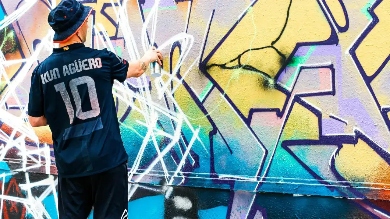 Mann mit Hut und Trikot sprüht mit Spraydose auf bunte Graffiti-Wand.