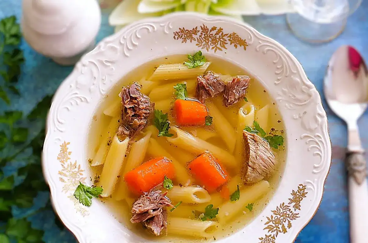 Klarowny rosół z jagnięciny z makaronem penne, kawałkami mięsa i marchewką, ozdobiony natką pietruszki.