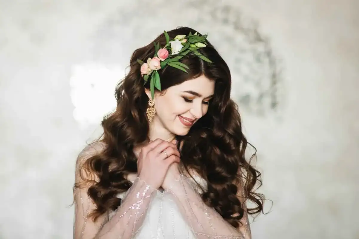Uśmiechnięta panna młoda z delikatnymi lokami na wesele, ozdobiona wiankiem z kwiatów i liści.