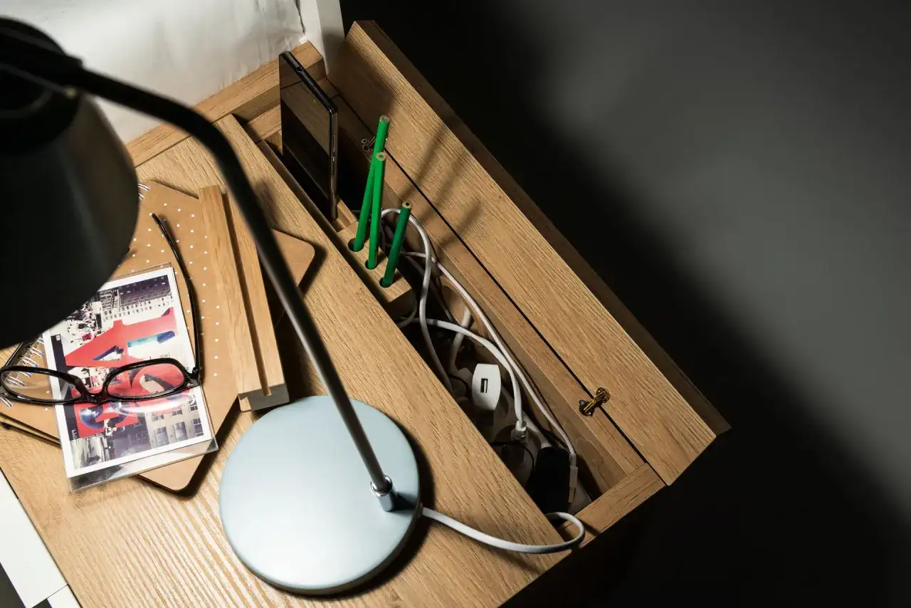 Drewniana szafka nocna z przegródkami na telefon i zielone ołówki. W schowku na kable panuje porządek dzięki praktycznym zabezpieczeniom na kable.