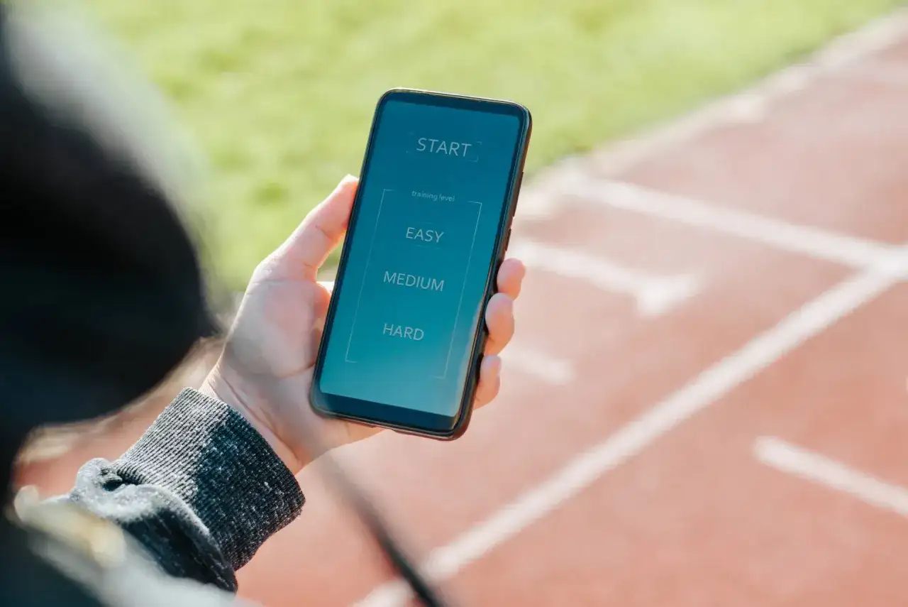 Dłoń trzyma smartfon z ekranem aplikacji do biegania. Wybierz poziom treningu: łatwy, średni lub trudny. To najlepsza aplikacja do biegania!