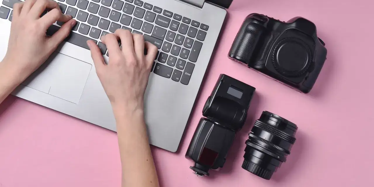 Ręce piszą na laptopie, obok aparat, lampa błyskowa i obiektyw. Szukasz, gdzie sprzedać aparat fotograficzny?
