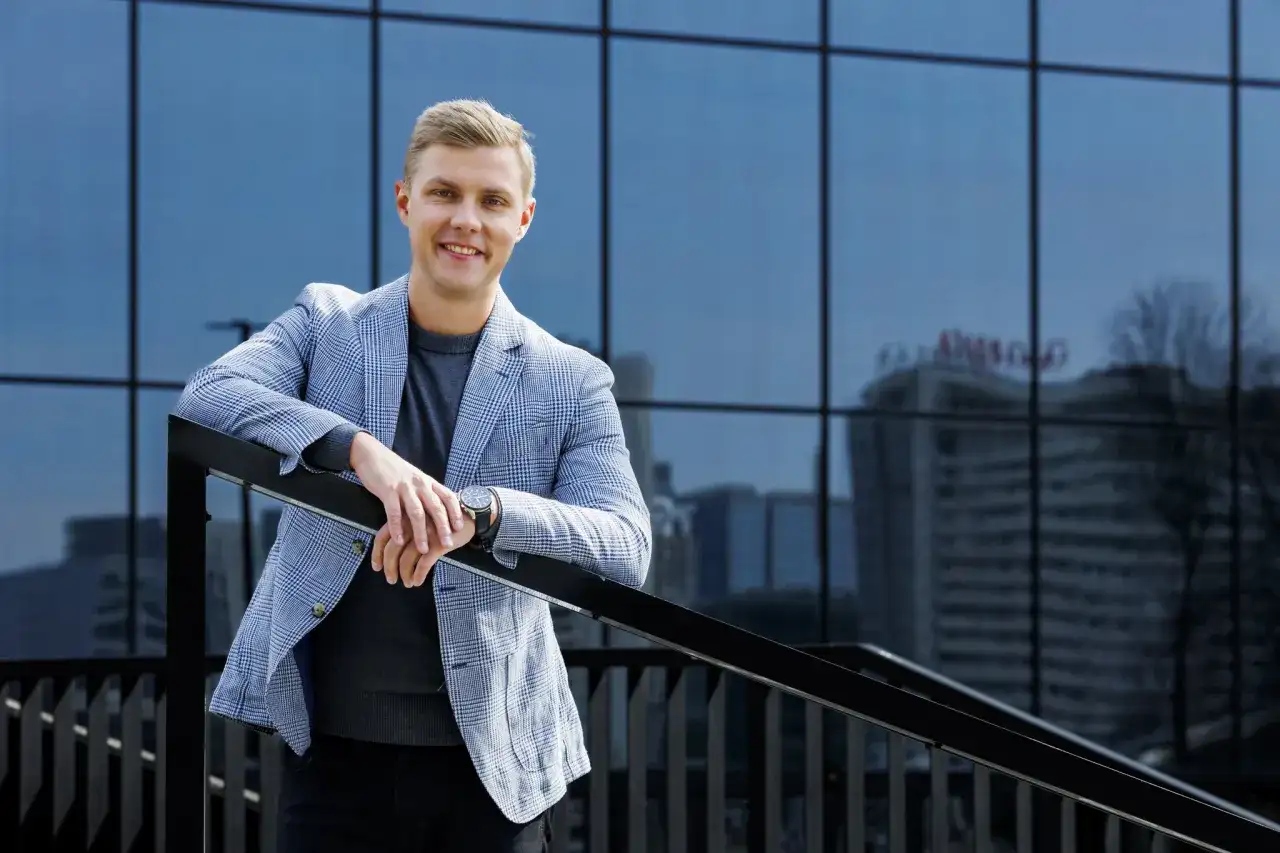 Uśmiechnięty mężczyzna w marynarce w kratę podczas sesji biznesowej w plenerze, oparty o balustradę na tle nowoczesnego budynku.