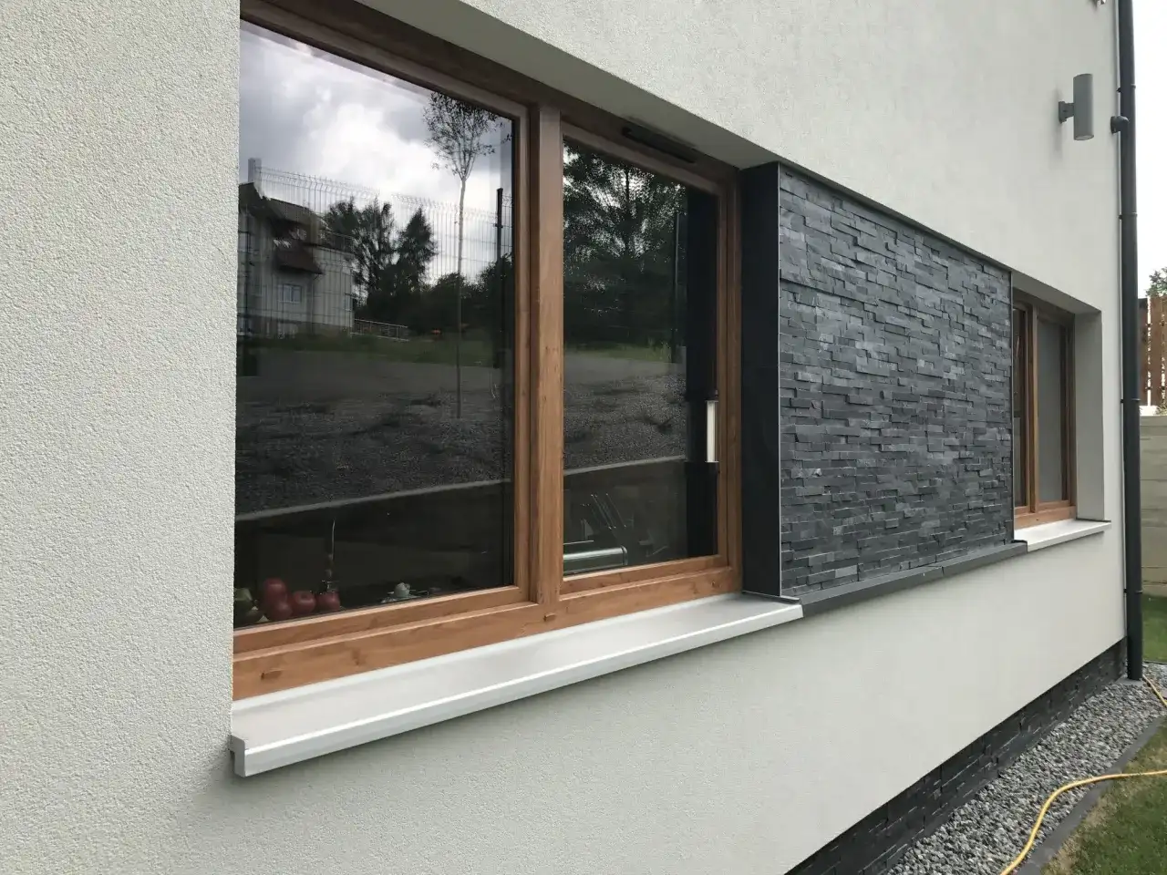 Nowoczesne okna z drewnianymi ramami i kamiennymi opaskami wokół okien, odbijające niebo i otoczenie.