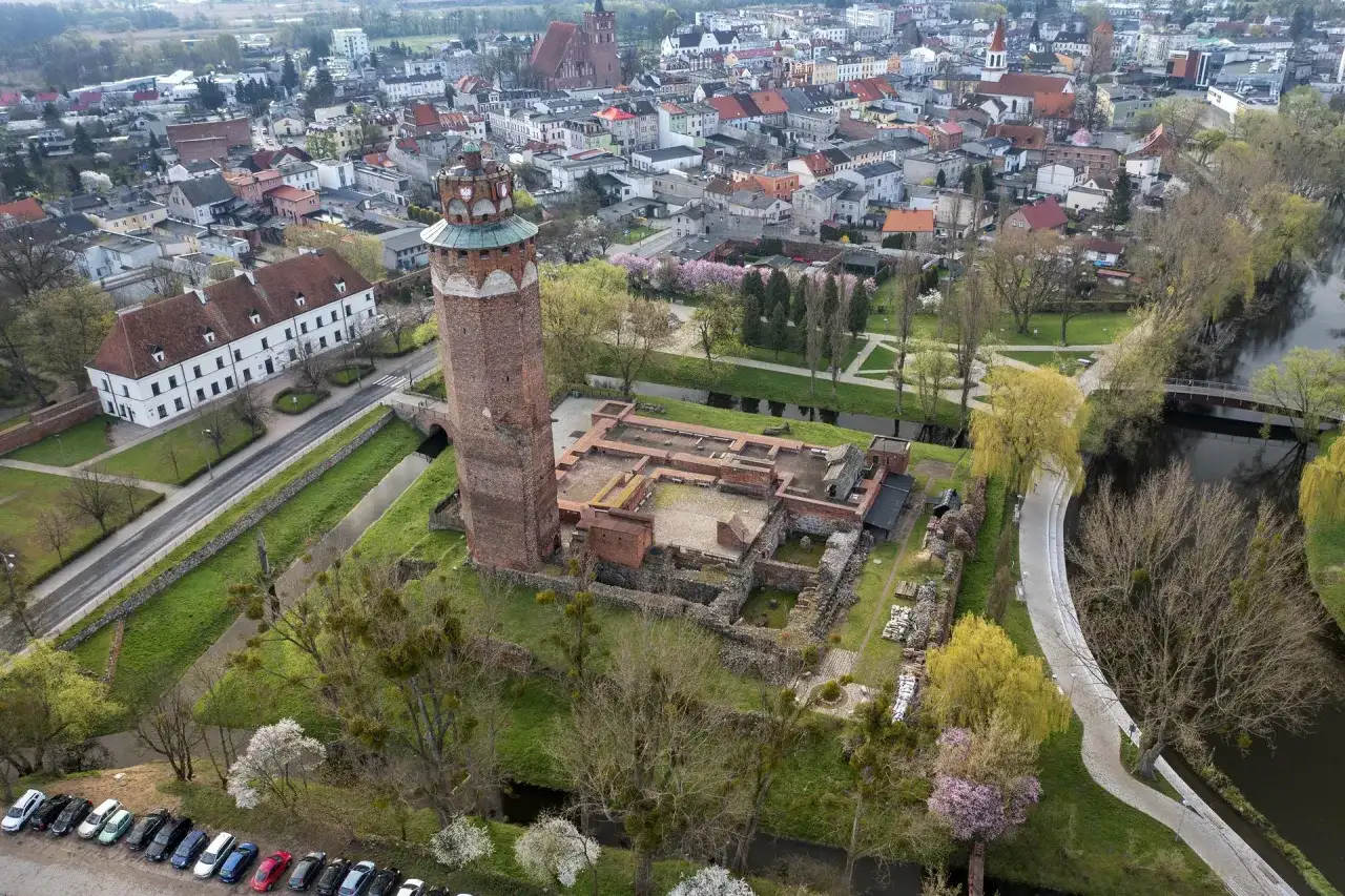 Ruiny zamku krzyżackiego w Brodnicy z charakterystyczną wieżą, otoczone zielenią i wodą, w tle rozciąga się miasto.