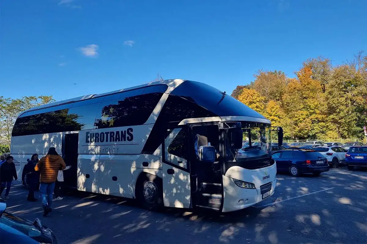 Biały autokar firmy Eurotrans z otwartymi drzwiami, zaparkowany na tle jesiennych drzew i innych samochodów.