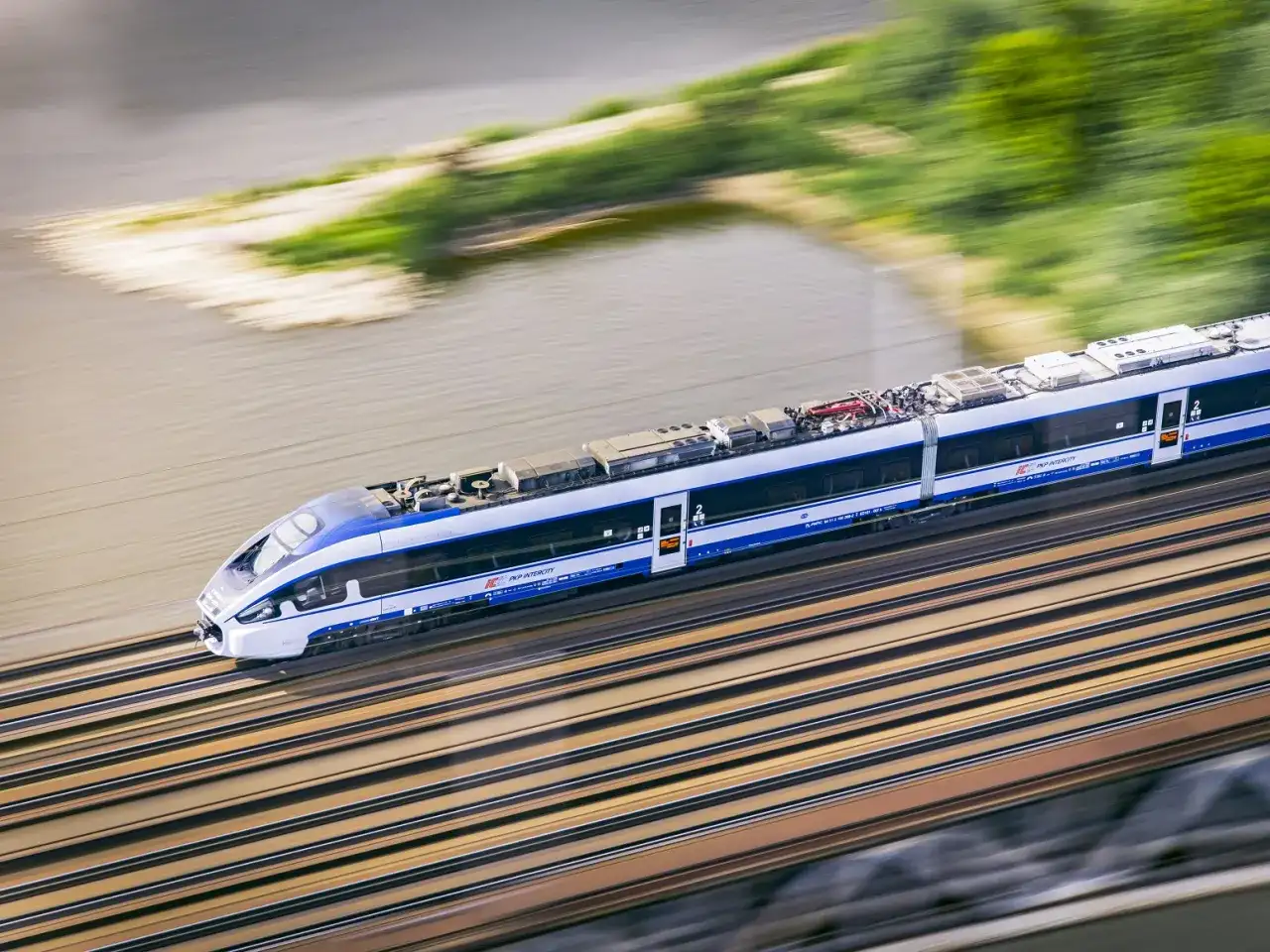Pędzący pociąg Intercity w ruchu, z rozmytym tłem rzeki i zieleni.