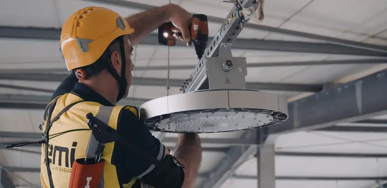 Pracownik w żółtym kasku i kamizelce montuje nowoczesne oświetlenie LED. To wymiana oświetlenia na LED – remont czy modernizacja hali.