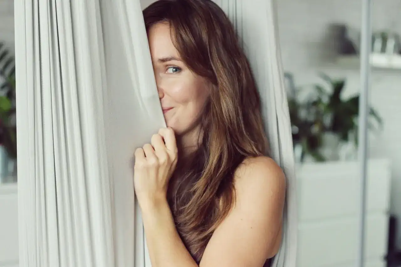 Uśmiechnięta kobieta z brązowymi włosami, ukrywająca się za zasłoną. Jej spojrzenie mówi "jestem jaka jestem cytaty".