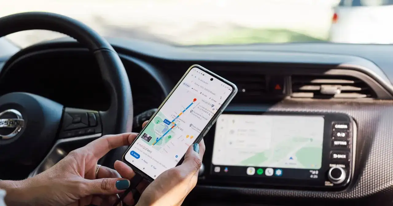 Dłoń trzyma telefon z nawigacją GPS w samochodzie, obok kierownicy.