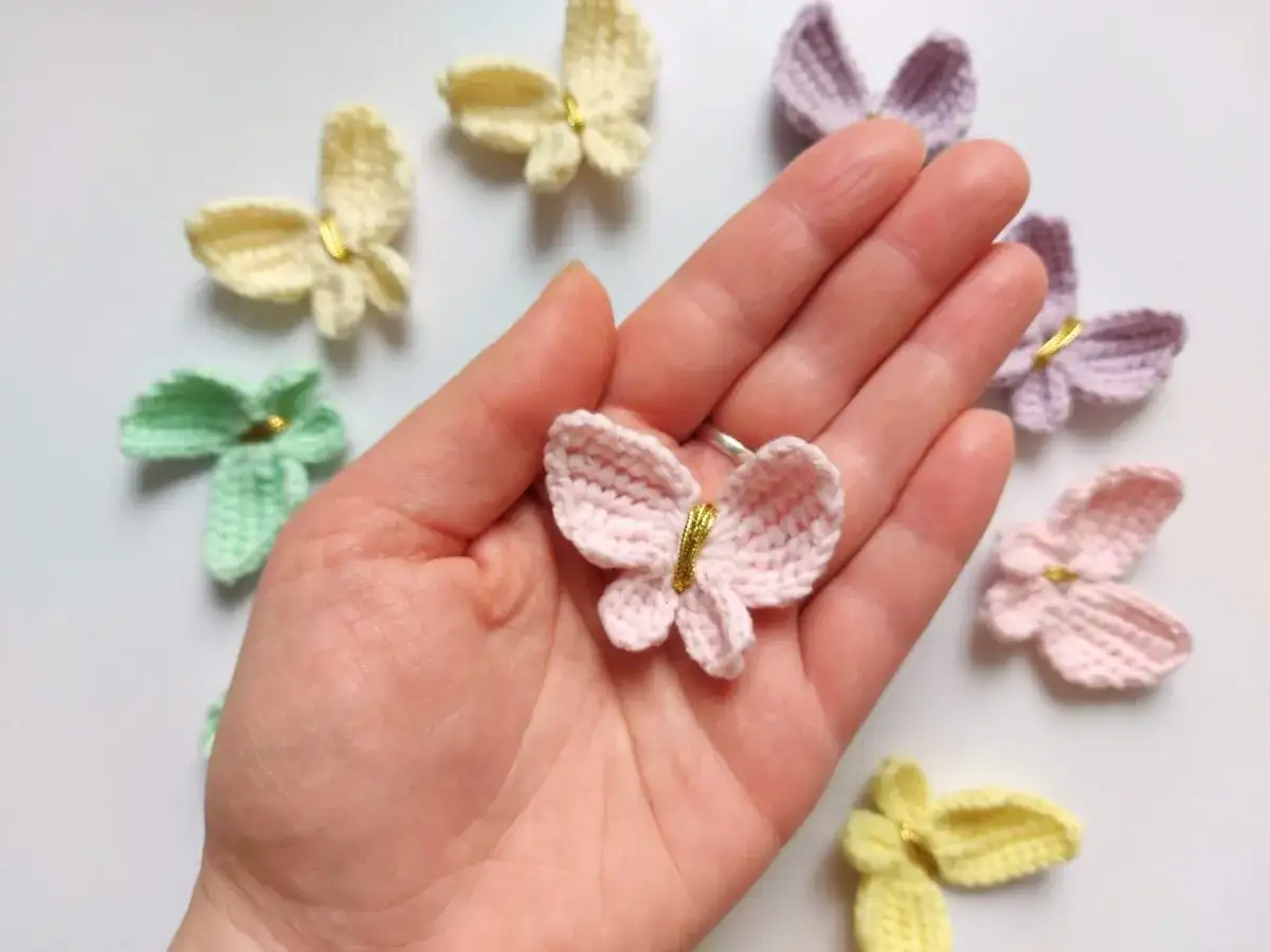 Różowy motylek na szydełku w dłoni, otoczony innymi kolorowymi motylkami.