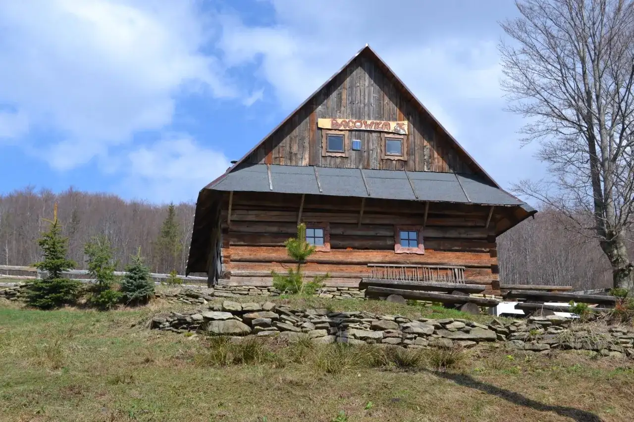 Drewniana bacówka w górach z napisem "BACÓWKA" na dachu, otoczona drzewami i kamiennym murkiem.
